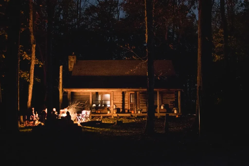 A group of people sit around a campfire in front of a warmly lit wooden cabin surrounded by trees at night.
